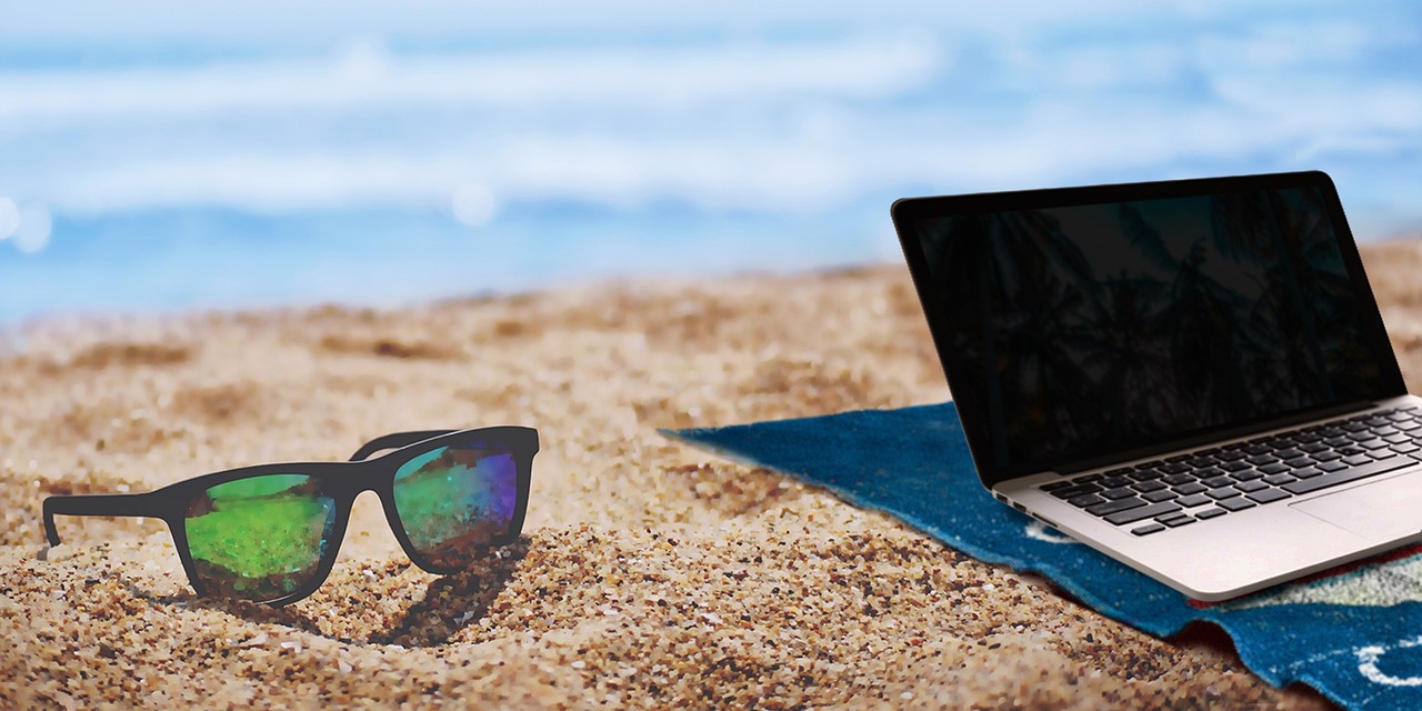 a laptop sitting open on a beach blanket on the beach sand next to a pair of sunglasses.